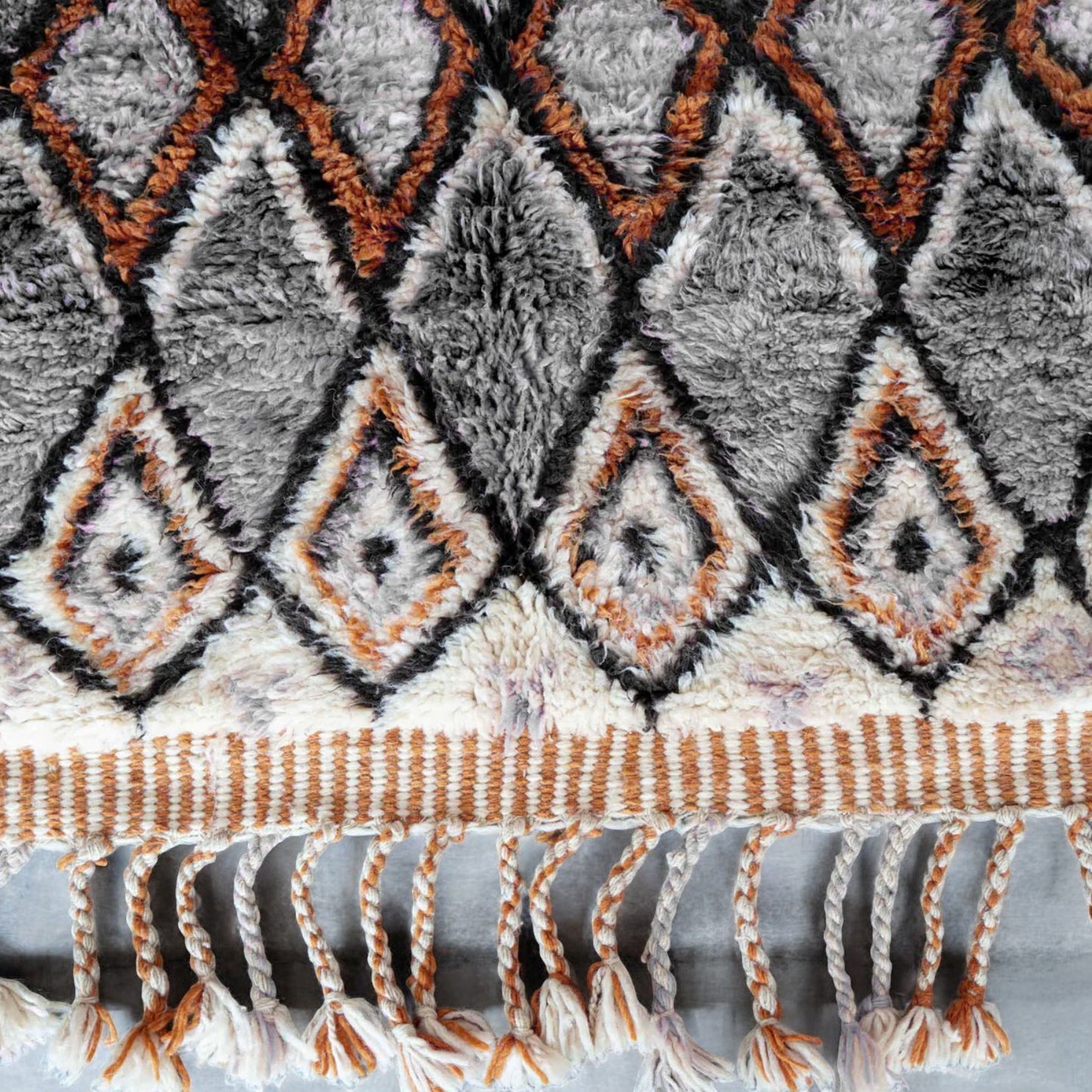 A handmade Moroccan wool rug featuring a sophisticated geometric diamond pattern in neutral tones, orange, and black, accented with intricate details and tassels on the edges. This rug blends traditional Berber craftsmanship with modern interior design styles.