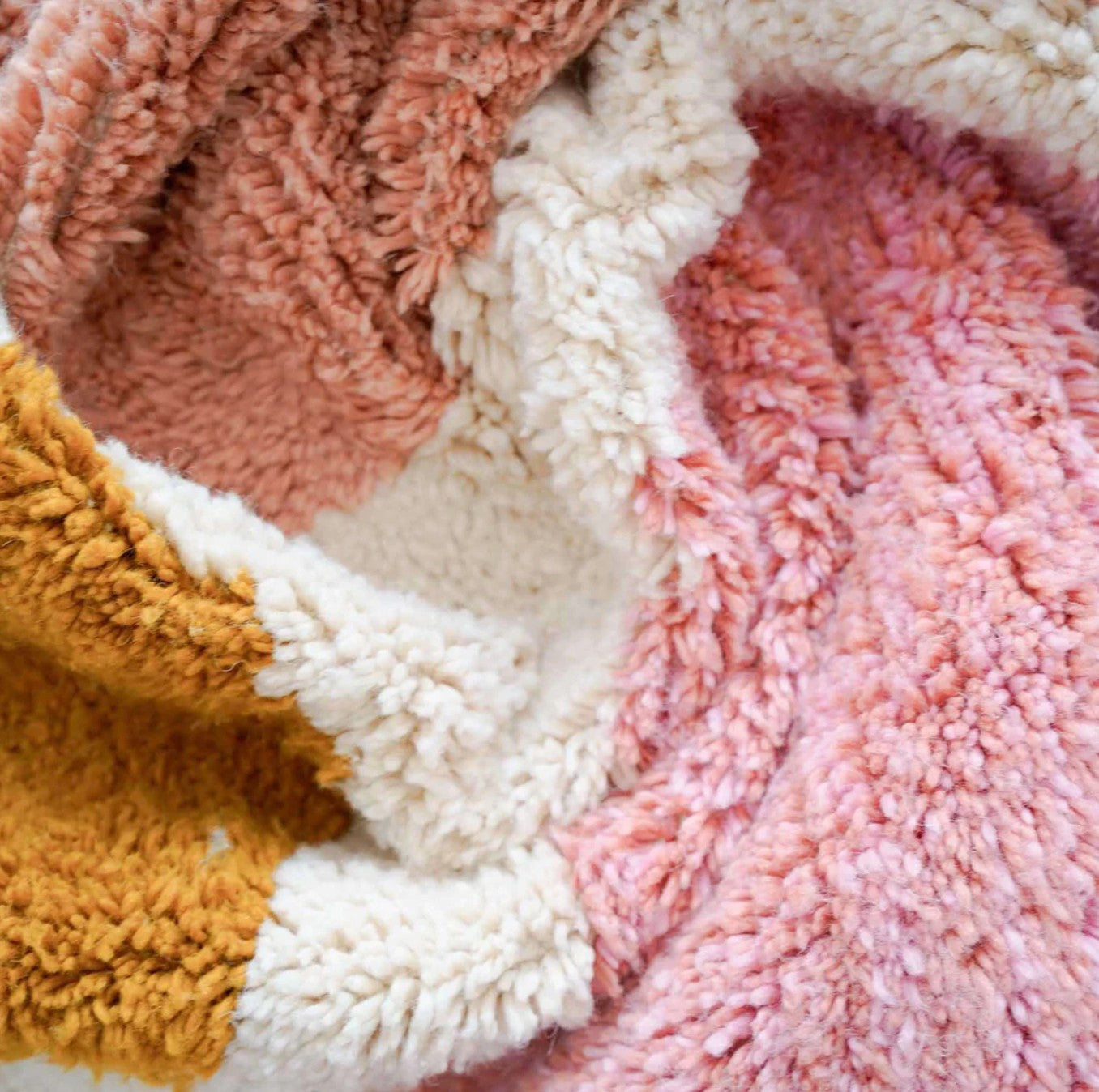 A handmade Moroccan wool rug featuring a vibrant color block design with shades of pink, blue, mustard yellow, and neutral tones, accented by tassels along the edge. Displayed against a bright yellow wall with natural green and wooden decor elements.