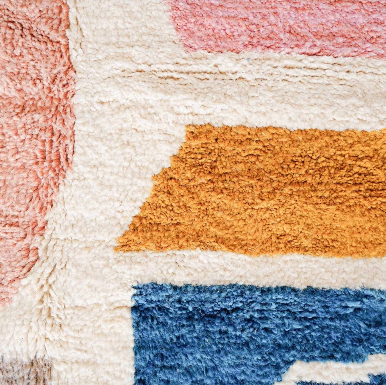 A handmade Moroccan wool rug featuring a vibrant color block design with shades of pink, blue, mustard yellow, and neutral tones, accented by tassels along the edge. Displayed against a bright yellow wall with natural green and wooden decor elements.