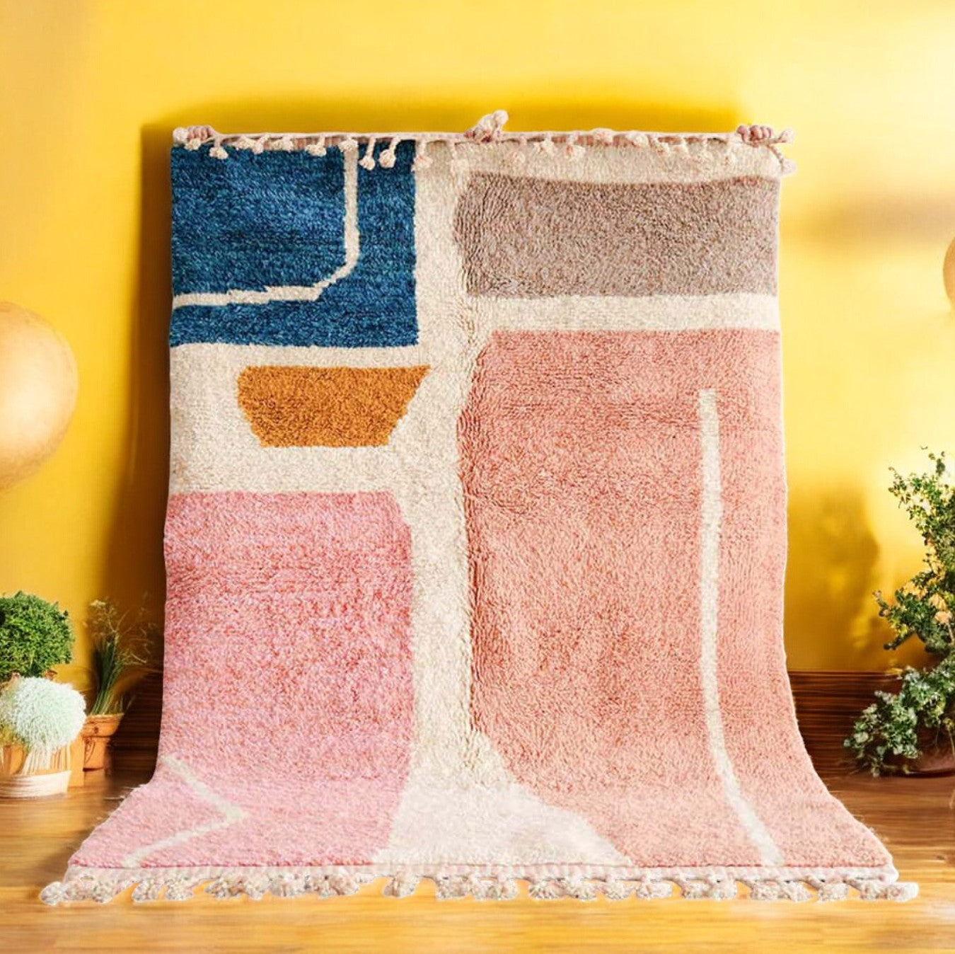 A handmade Moroccan wool rug featuring a vibrant color block design with shades of pink, blue, mustard yellow, and neutral tones, accented by tassels along the edge. Displayed against a bright yellow wall with natural green and wooden decor elements.