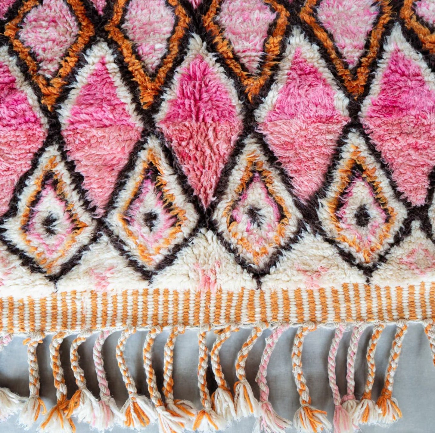 A handmade Moroccan wool rug featuring a vibrant geometric diamond pattern in shades of pink, orange, and black, with intricate detailing and tassels on the edges. A perfect blend of traditional Berber artistry and modern design aesthetics.