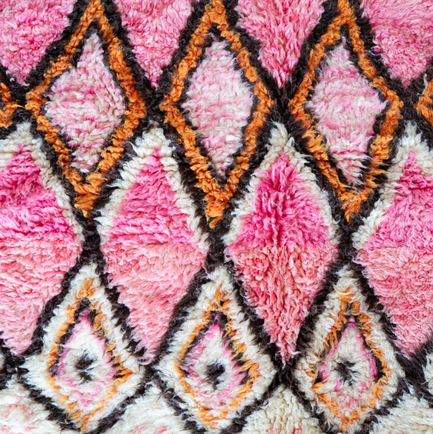 A handmade Moroccan wool rug featuring a vibrant geometric diamond pattern in shades of pink, orange, and black, with intricate detailing and tassels on the edges. A perfect blend of traditional Berber artistry and modern design aesthetics.