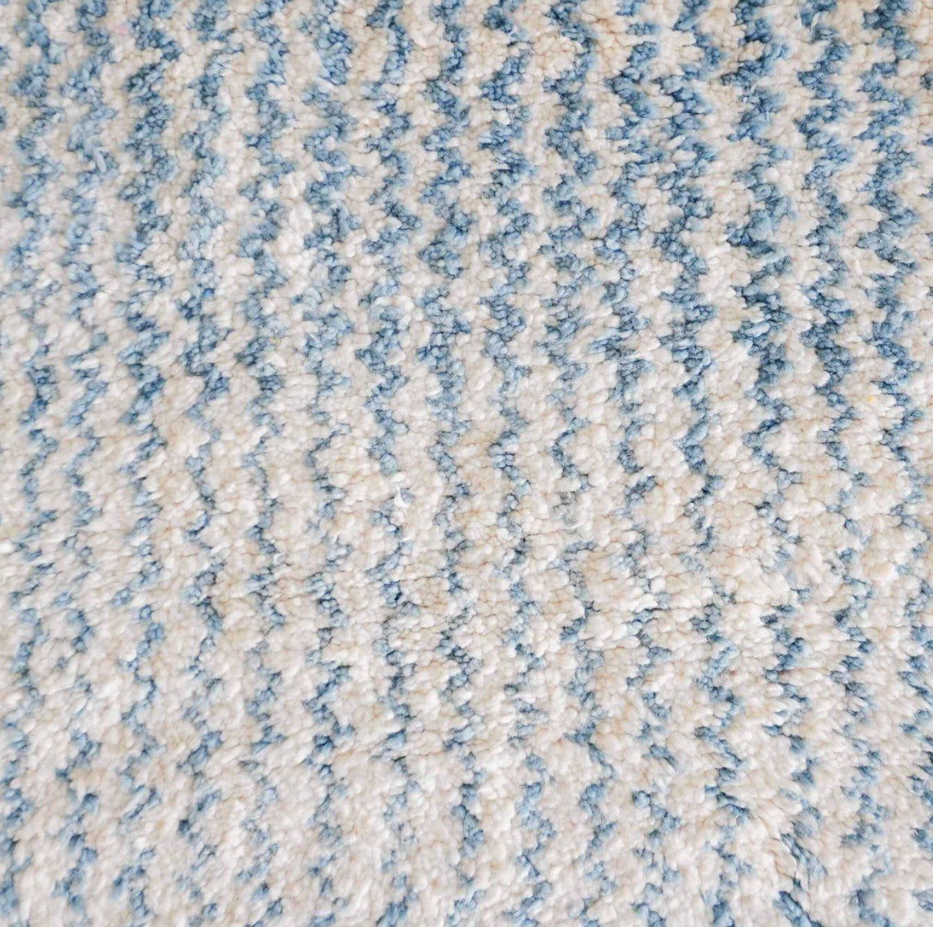 Handmade wool rug featuring a soothing sky blue and white color palette with intricate geometric patterns, accented by delicate fringe detailing, displayed on a warm wooden floor.