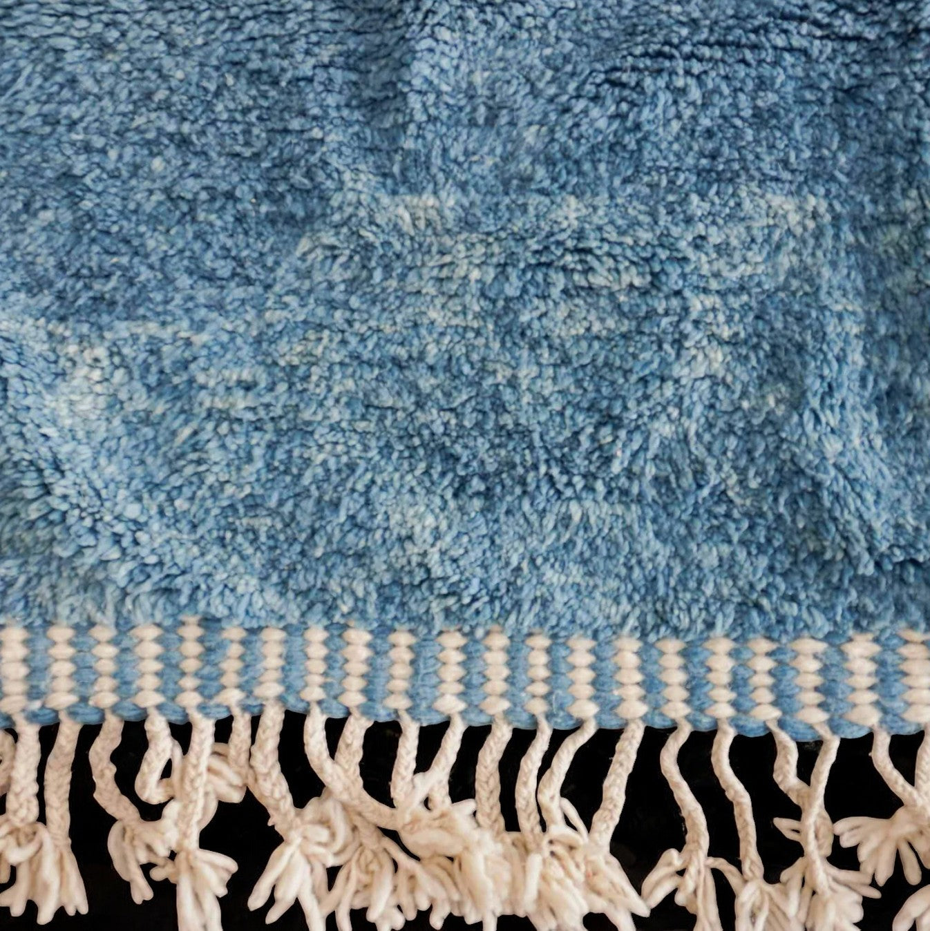 Handmade wool rug featuring a soothing sky blue and white color palette with intricate geometric patterns, accented by delicate fringe detailing, displayed on a warm wooden floor.