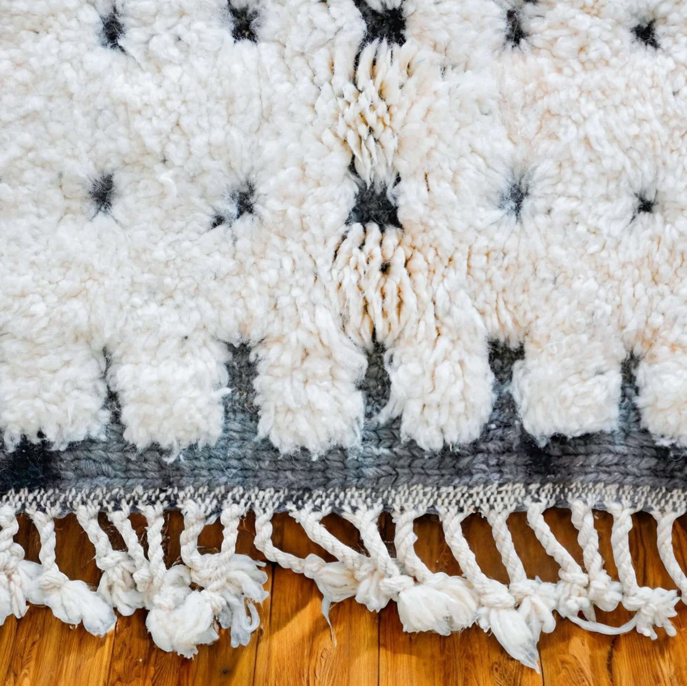Artisanal handmade wool rug in white with a dotted black stripe pattern and delicate fringed edges, displayed on a warm wooden floor, ideal for minimalist and modern decor.