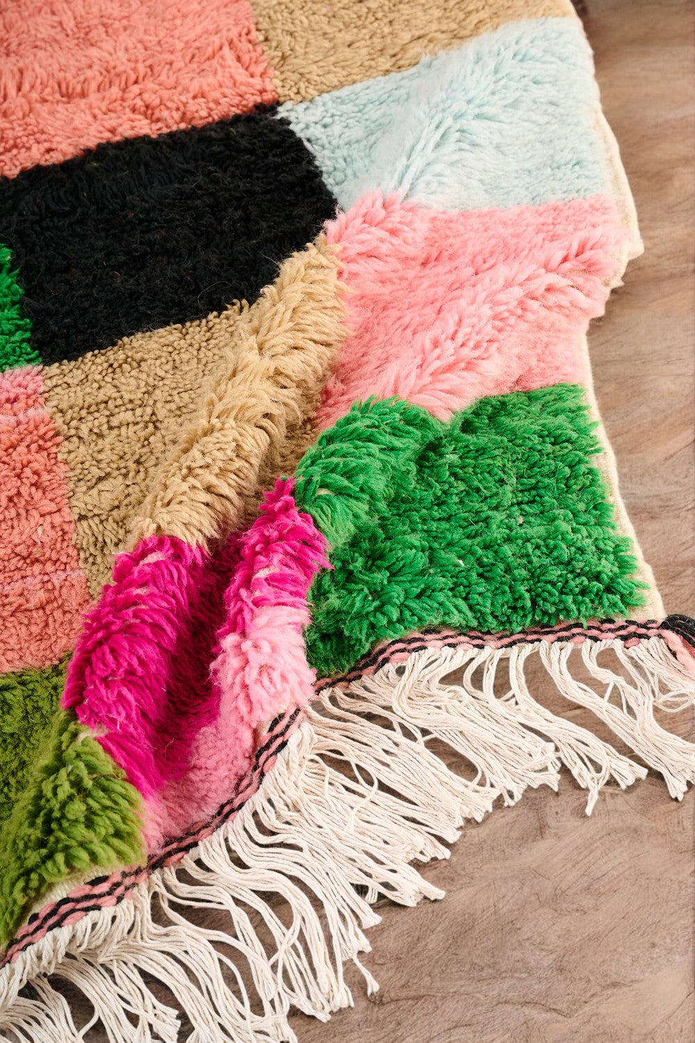 Handmade Moroccan wool rug featuring a bold multicolor checkerboard design with vibrant shades of pink, green, orange, black, beige, and blue, displayed on a herringbone wooden floor in a modern boho room with a rattan chair, potted plant, and minimalist wall art