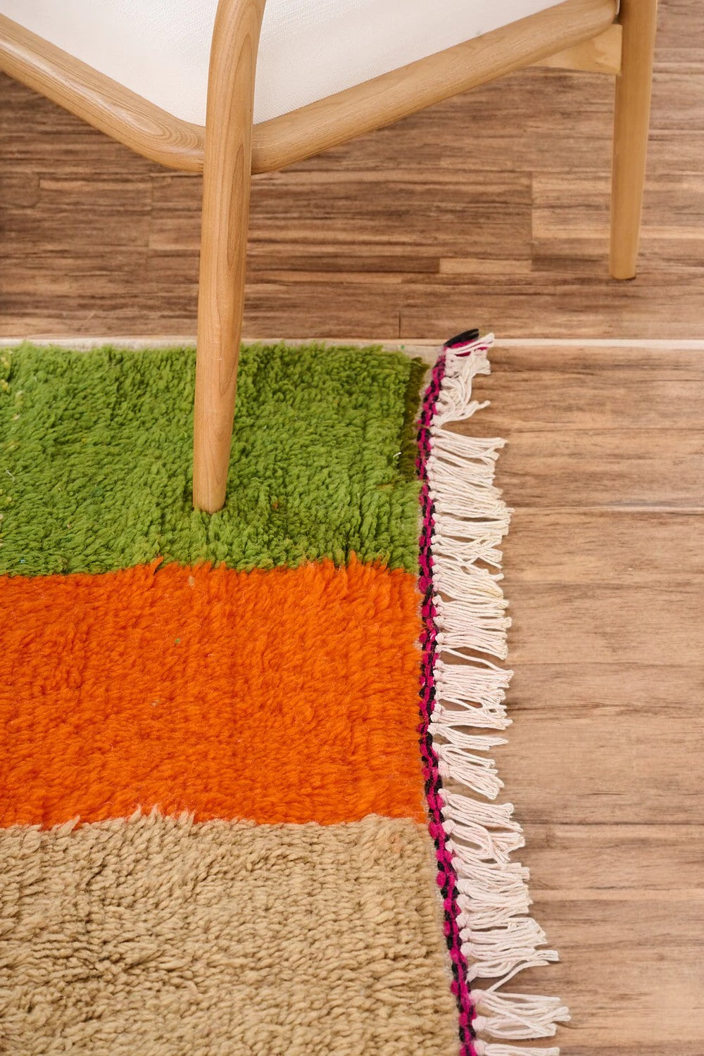 Handmade Moroccan wool rug featuring a bold multicolor checkerboard design with vibrant shades of pink, green, orange, black, beige, and blue, displayed on a herringbone wooden floor in a modern boho room with a rattan chair, potted plant, and minimalist wall art