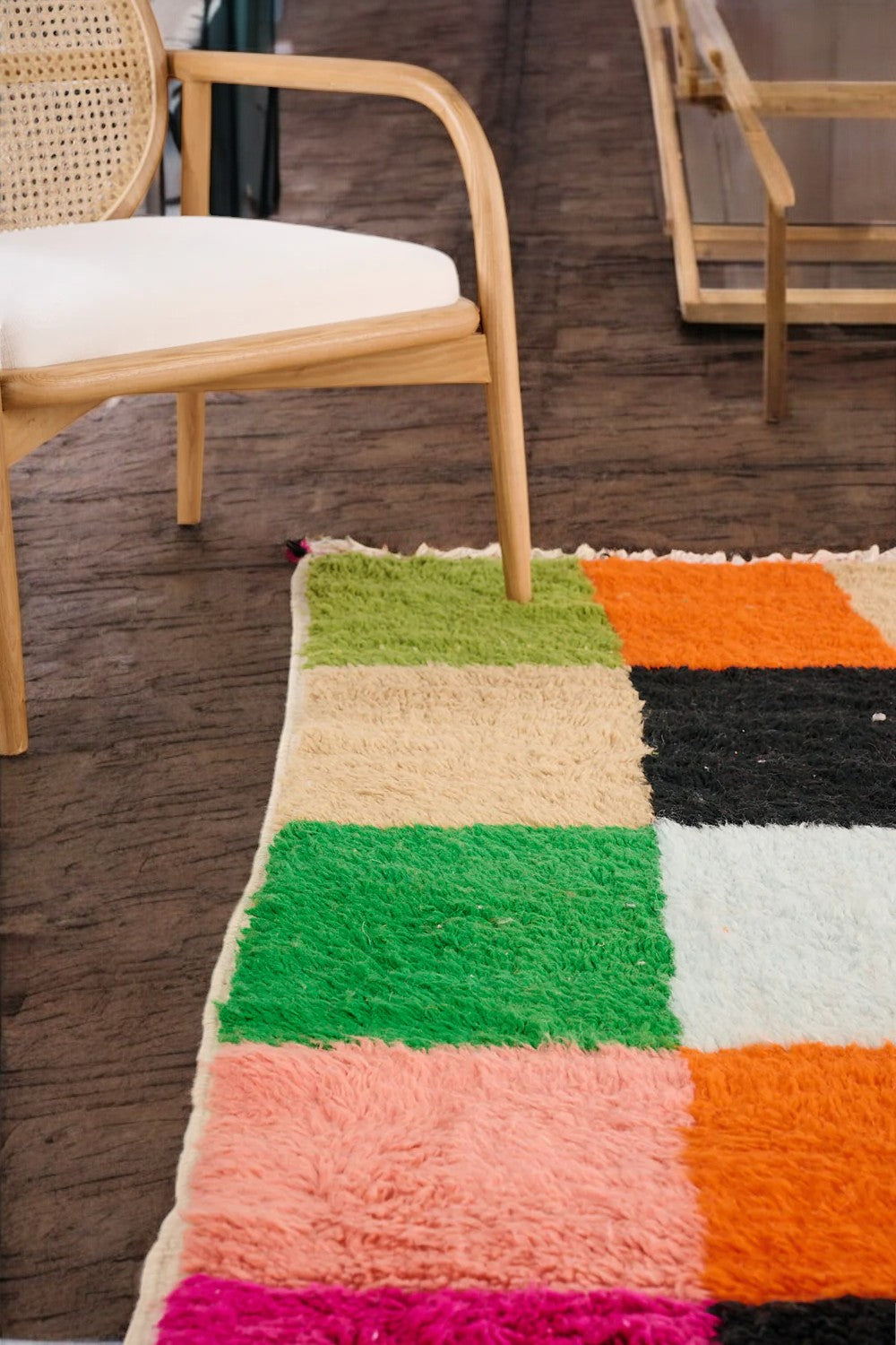 Handmade Moroccan wool rug featuring a bold multicolor checkerboard design with vibrant shades of pink, green, orange, black, beige, and blue, displayed on a herringbone wooden floor in a modern boho room with a rattan chair, potted plant, and minimalist wall art