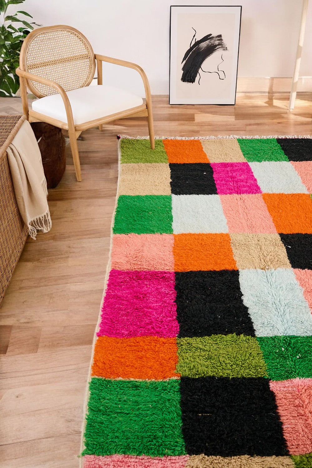 Handmade Moroccan wool rug featuring a bold multicolor checkerboard design with vibrant shades of pink, green, orange, black, beige, and blue, displayed on a herringbone wooden floor in a modern boho room with a rattan chair, potted plant, and minimalist wall art