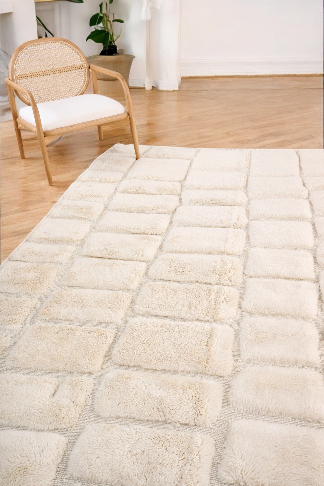 Handmade Moroccan wool rug in a soft cream color featuring a textured square pattern, displayed on a wooden floor in a bright minimalist room with a rattan chair, potted plant, and black-and-white abstract wall art.