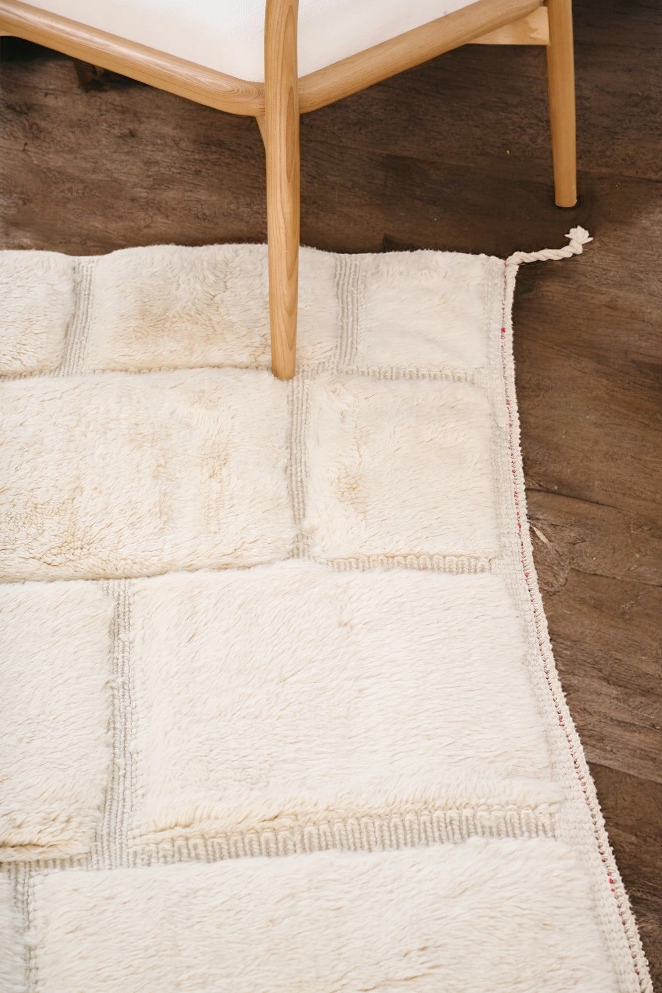 Handmade Moroccan wool rug in a soft cream color featuring a textured square pattern, displayed on a wooden floor in a bright minimalist room with a rattan chair, potted plant, and black-and-white abstract wall art.
