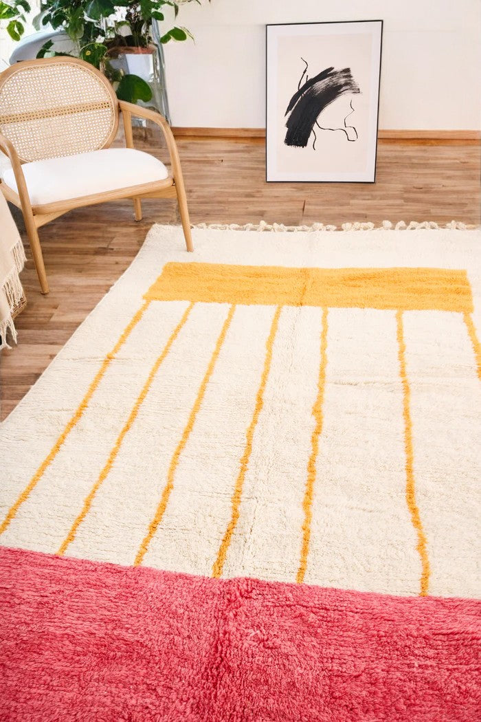 Handmade Moroccan wool rug with a minimalist cream base featuring vibrant yellow vertical stripes and a bold red border, styled on a wooden floor in a cozy boho room with a rattan chair, potted plant, and black-and-white abstract wall art
