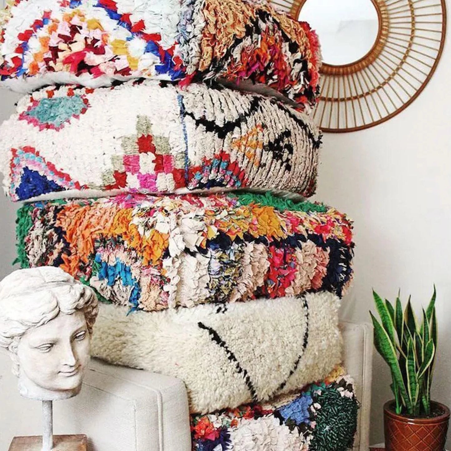 Stack of four colorful Moroccan-style floor cushions with intricate geometric patterns, vibrant reds, pinks, beige, and black tones, against a white brick wall background.
