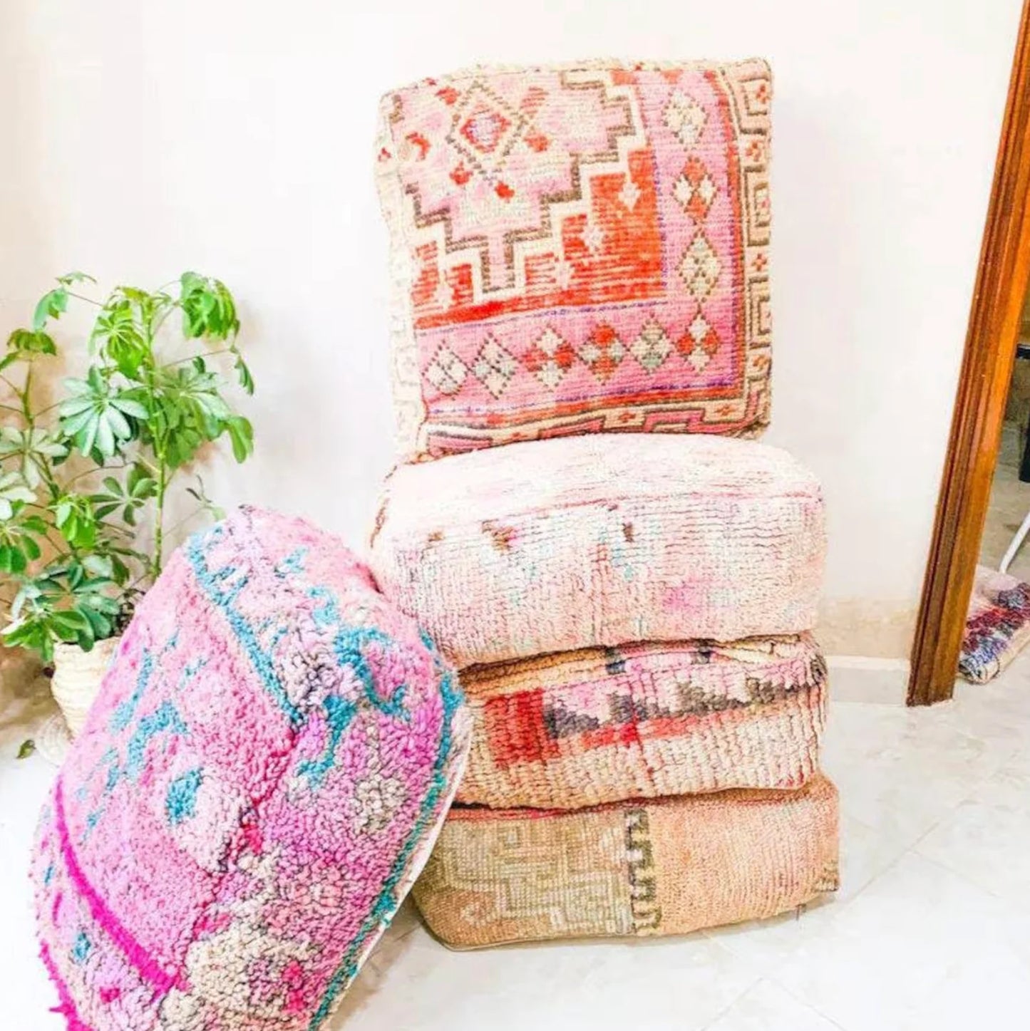 Stack of four colorful Moroccan-style floor cushions with intricate geometric patterns, vibrant reds, pinks, beige, and black tones, against a white brick wall background.