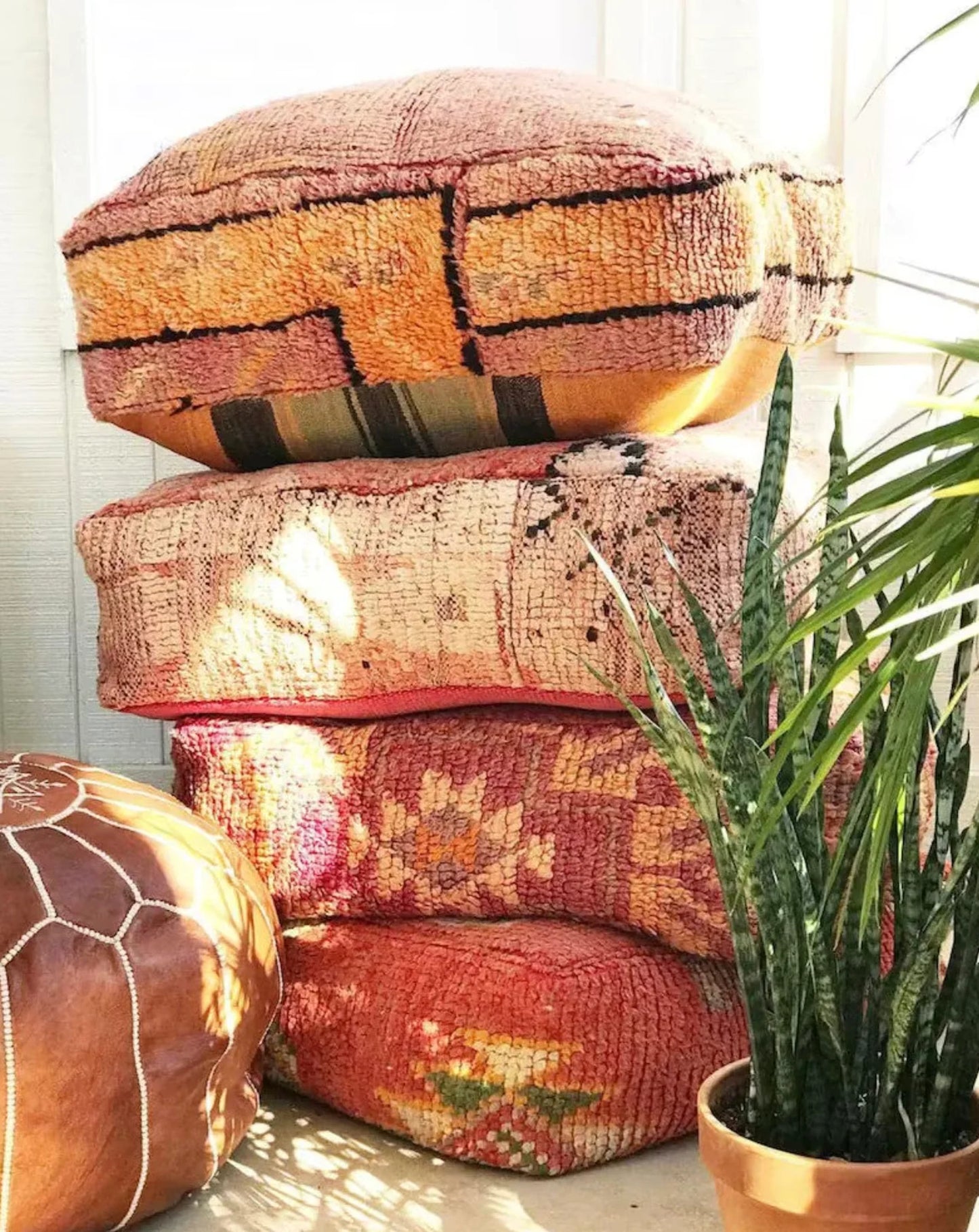 Stack of four colorful Moroccan-style floor cushions with intricate geometric patterns, vibrant reds, pinks, beige, and black tones, against a white brick wall background.