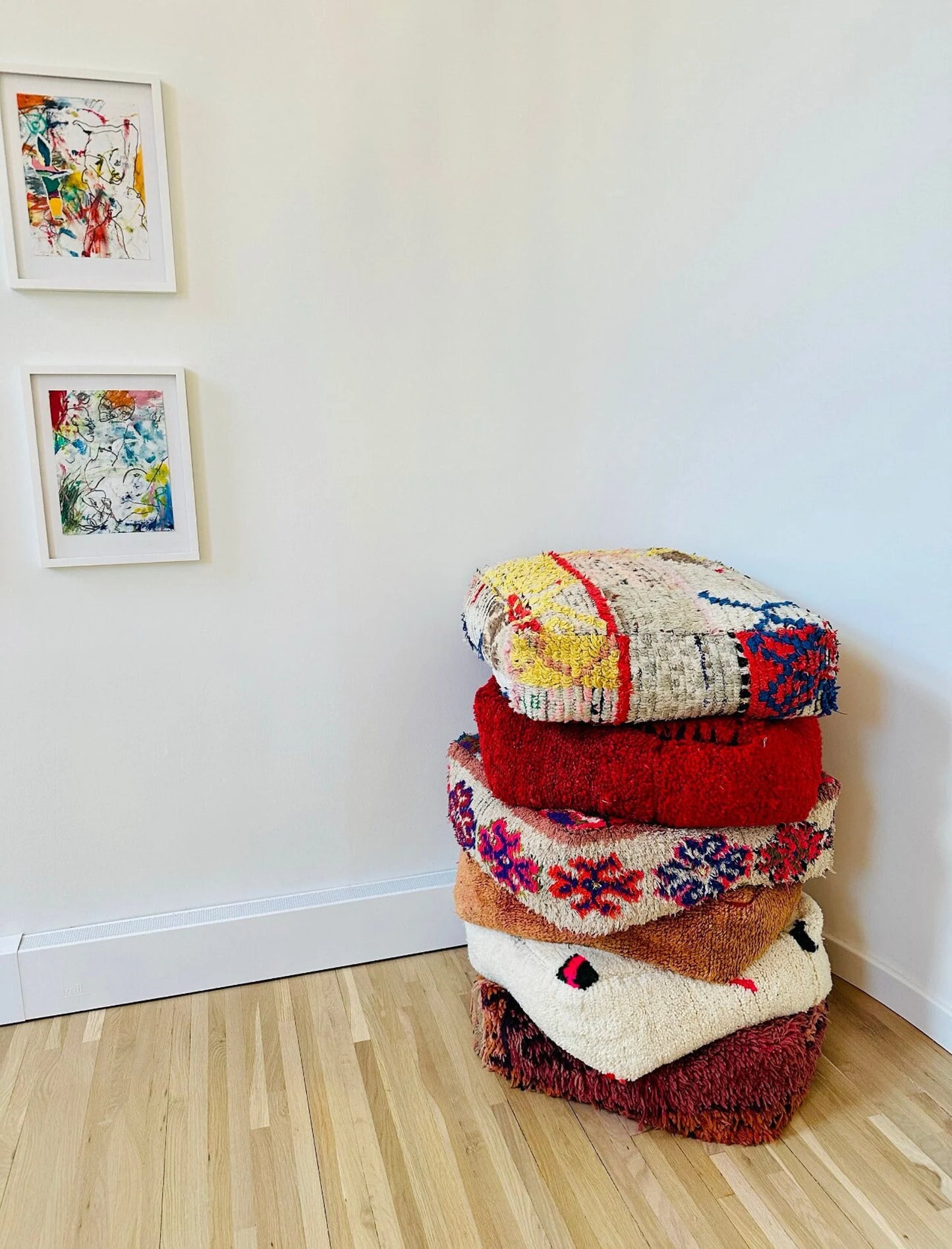 Stack of four colorful Moroccan-style floor cushions with intricate geometric patterns, vibrant reds, pinks, beige, and black tones, against a white brick wall background.