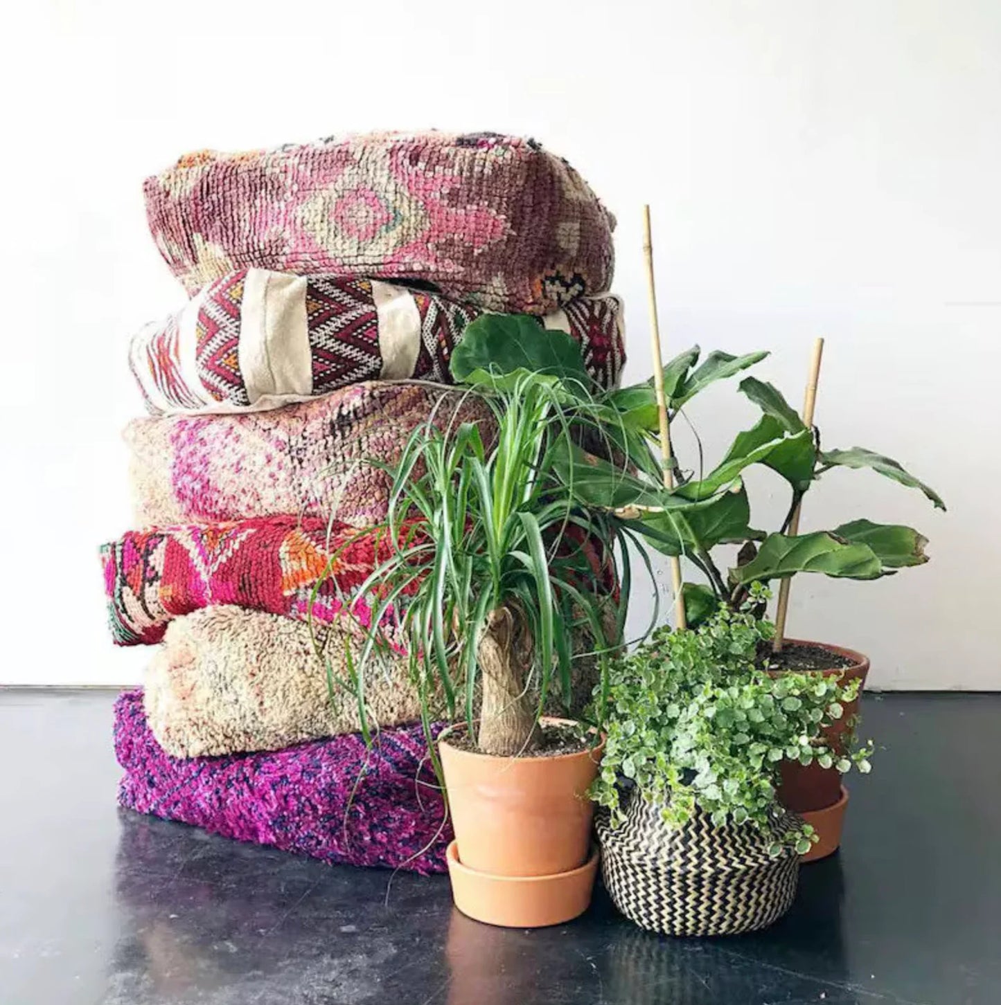 Stack of four colorful Moroccan-style floor cushions with intricate geometric patterns, vibrant reds, pinks, beige, and black tones, against a white brick wall background.