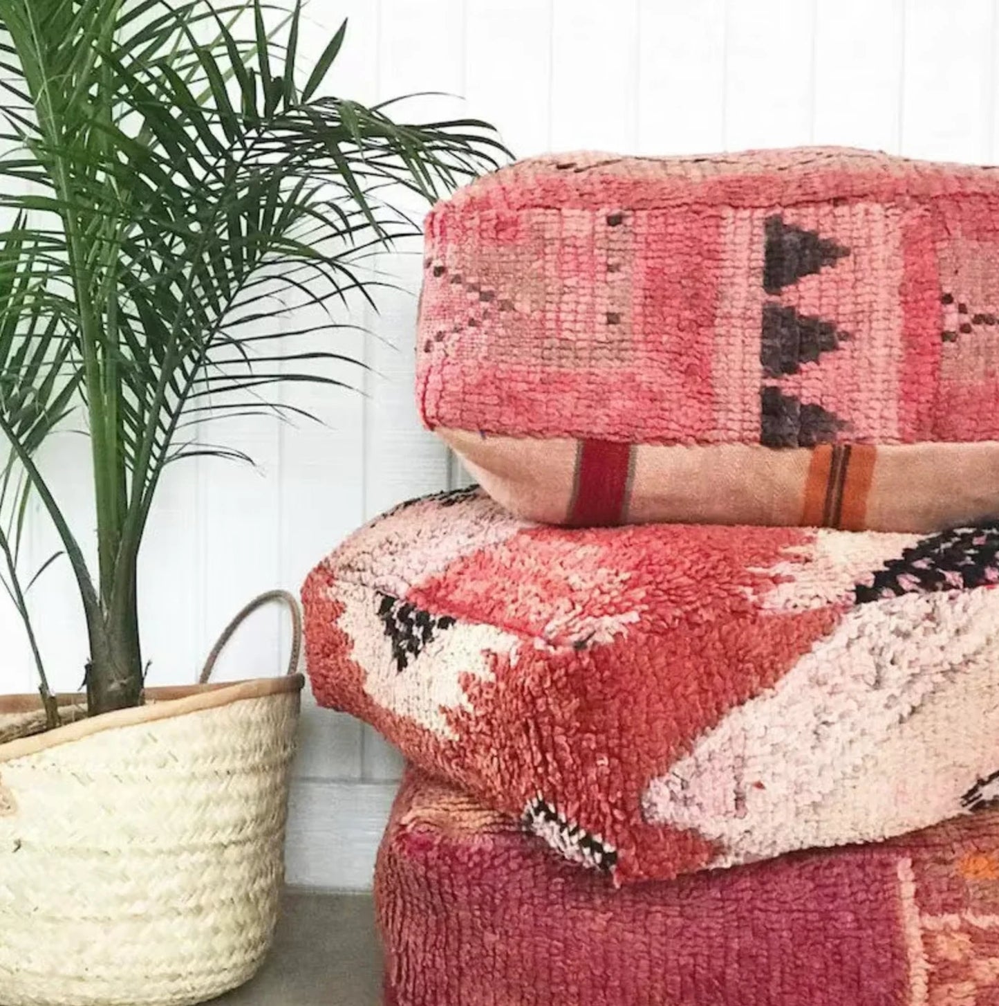 Stack of four colorful Moroccan-style floor cushions with intricate geometric patterns, vibrant reds, pinks, beige, and black tones, against a white brick wall background.