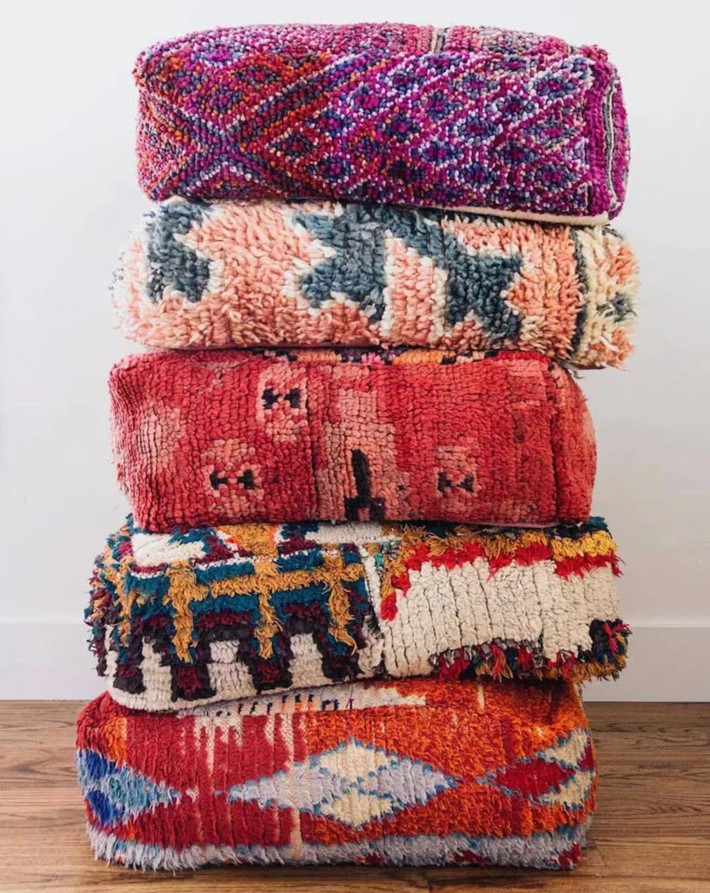 Stack of four colorful Moroccan-style floor cushions with intricate geometric patterns, vibrant reds, pinks, beige, and black tones, against a white brick wall background.