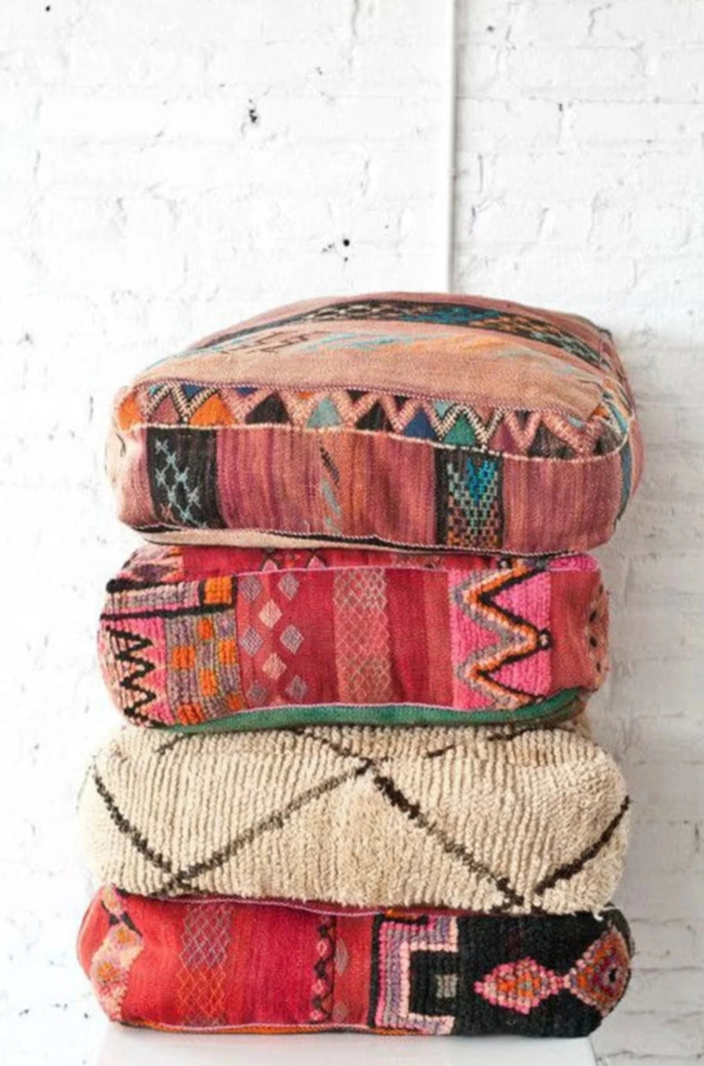 Stack of four colorful Moroccan-style floor cushions with intricate geometric patterns, vibrant reds, pinks, beige, and black tones, against a white brick wall background.