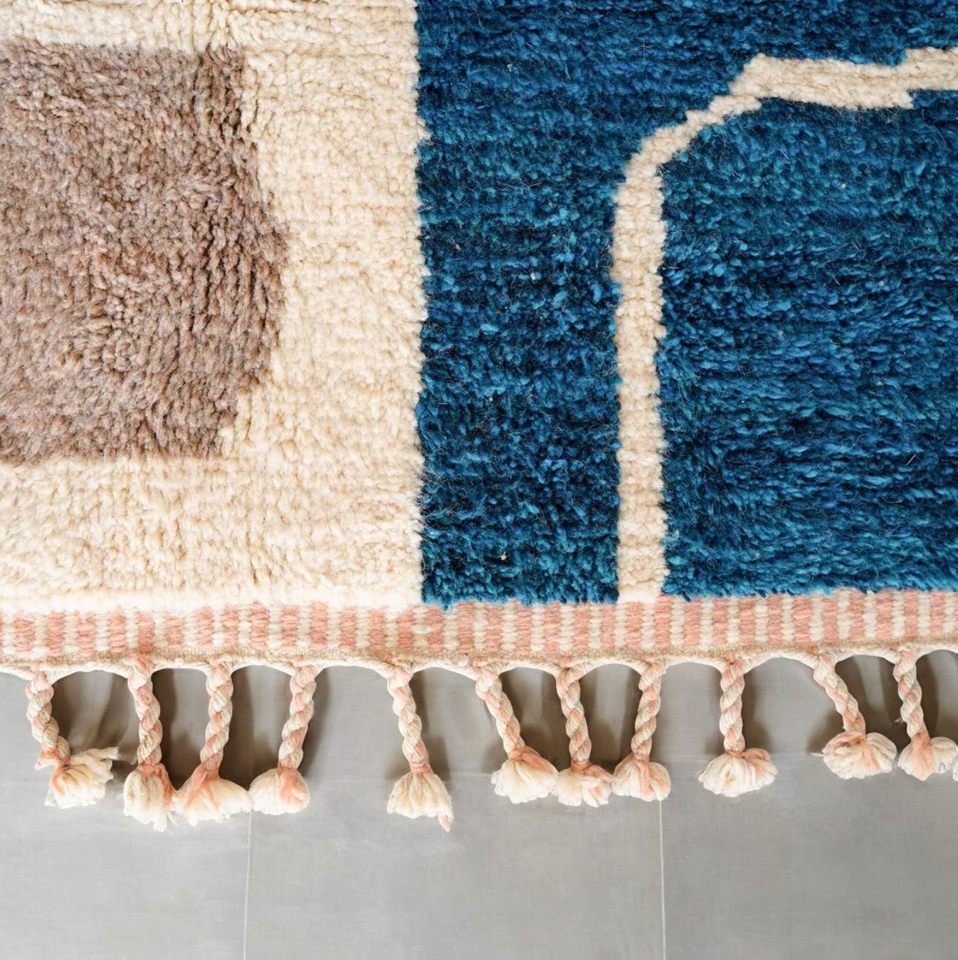A handmade Moroccan wool rug featuring a vibrant color block design with shades of pink, blue, mustard yellow, and neutral tones, accented by tassels along the edge. Displayed against a bright yellow wall with natural green and wooden decor elements.
