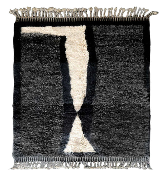 A large handmade wool rug featuring a bold black and beige geometric hourglass pattern, with intricate textures and fringed edges, beautifully styled on a wooden floor and set against a bright yellow wall with dramatic sunlight shadows.
