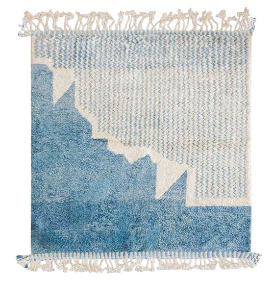 Handmade wool rug featuring a soothing sky blue and white color palette with intricate geometric patterns, accented by delicate fringe detailing, displayed on a warm wooden floor.