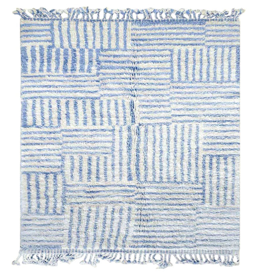 Artisanal handmade wool rug in a sky blue and white striped patchwork design, accented by delicate fringe, displayed on a natural wooden floor, ideal for coastal and modern decor.