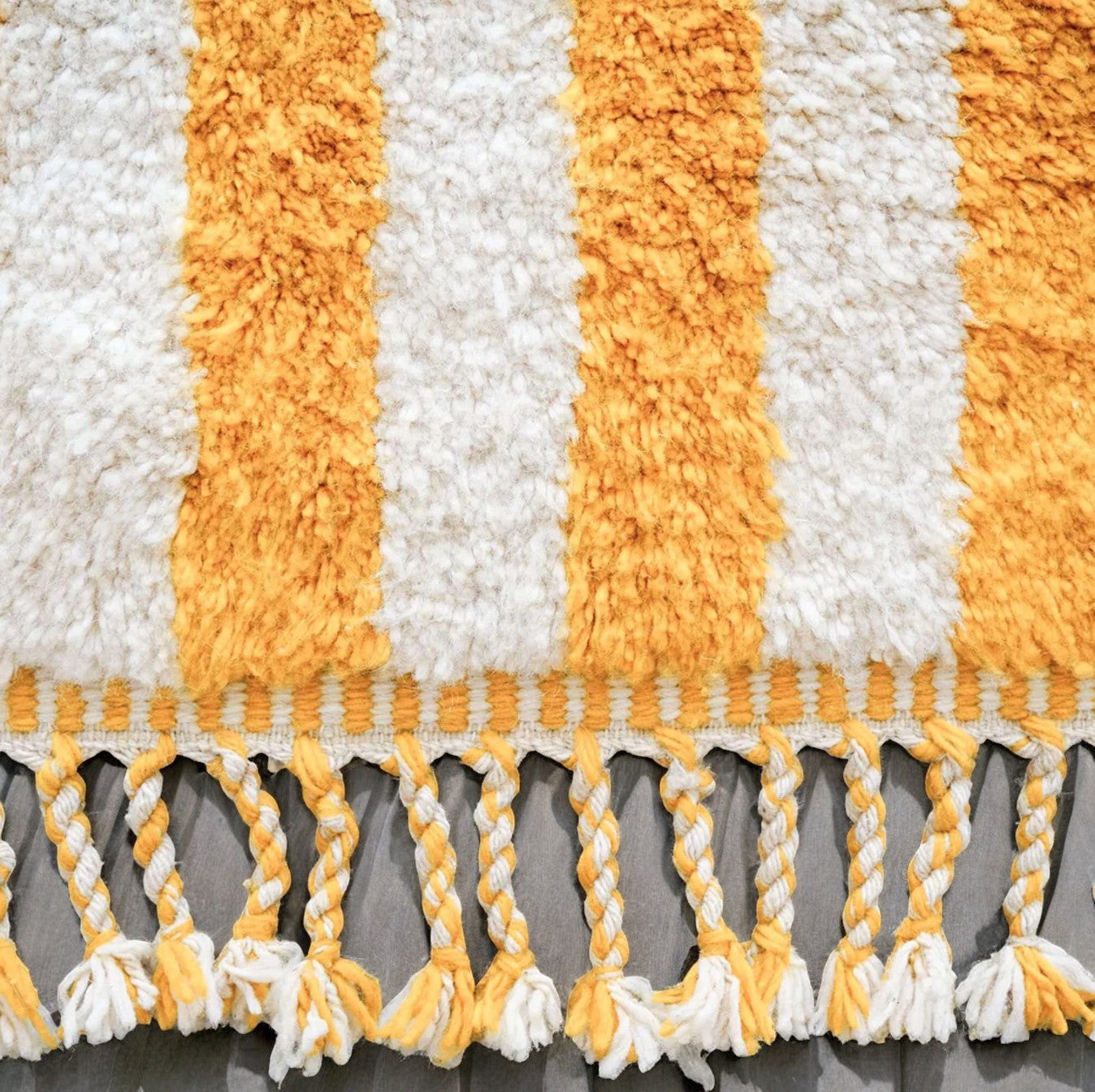 Artisanal handmade wool rug featuring bold yellow and white vertical stripes, accented by intricate fringe detailing, displayed on a neutral gray background, ideal for bright and cheerful modern decor.