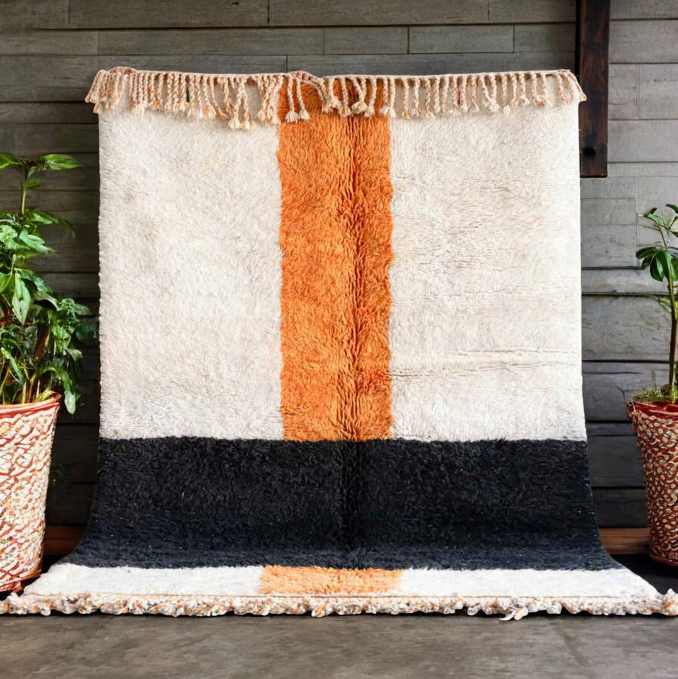 Handmade wool rug with bold horizontal black and vertical terracotta orange stripes on a soft white background, accented with intricate fringe along the edges, ideal for minimalist and modern decor styles."