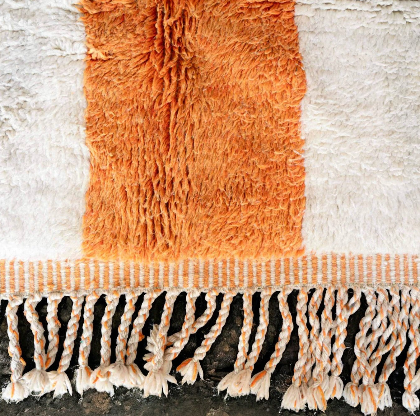 Handmade wool rug with bold horizontal black and vertical terracotta orange stripes on a soft white background, accented with intricate fringe along the edges, ideal for minimalist and modern decor styles."