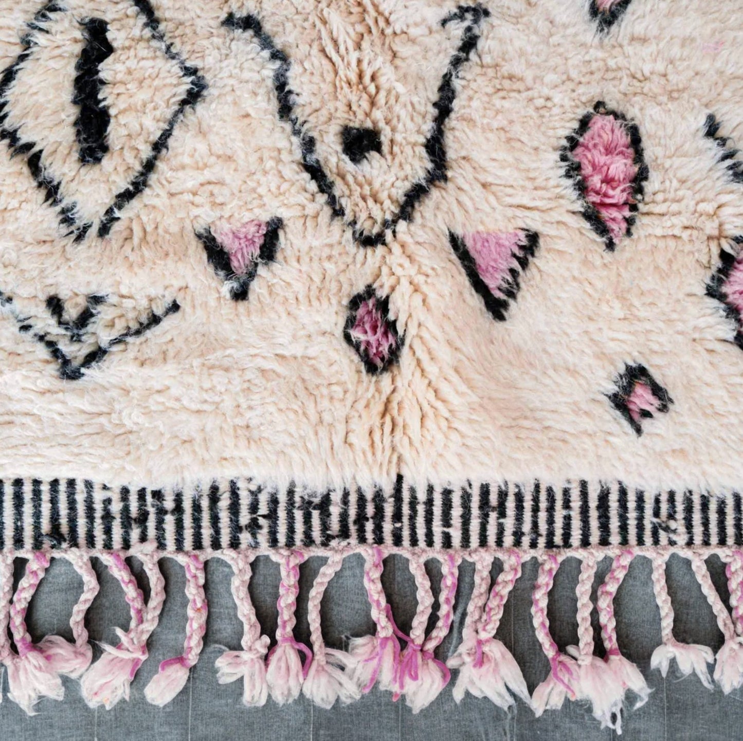 Handmade wool rug in soft white featuring pastel pink and black tribal geometric patterns, adorned with intricate fringe detailing, ideal for bohemian and modern feminine decor styles.