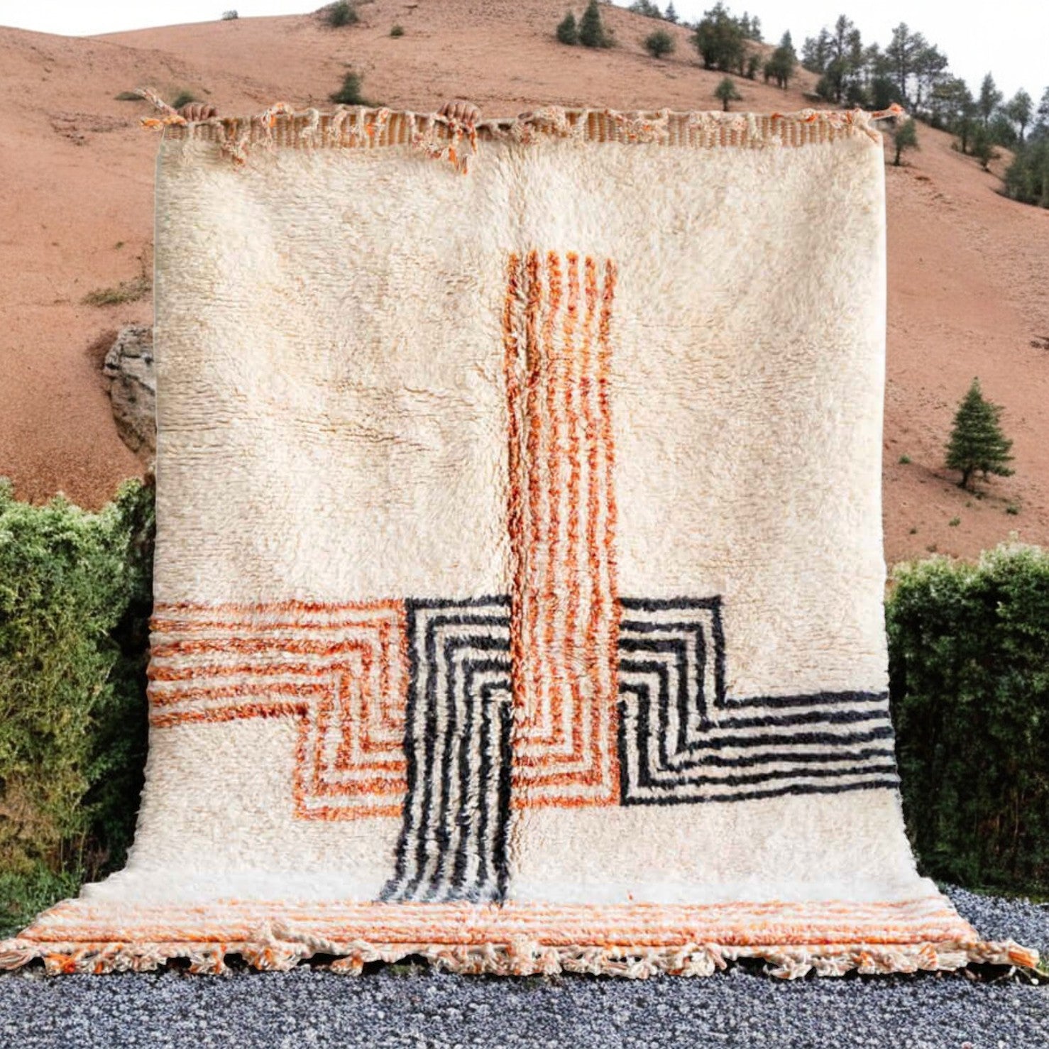 Artisanal handmade wool rug with vibrant orange and black geometric cross patterns on a soft white base, accented by intricate fringed edges, ideal for modern and eclectic decor.