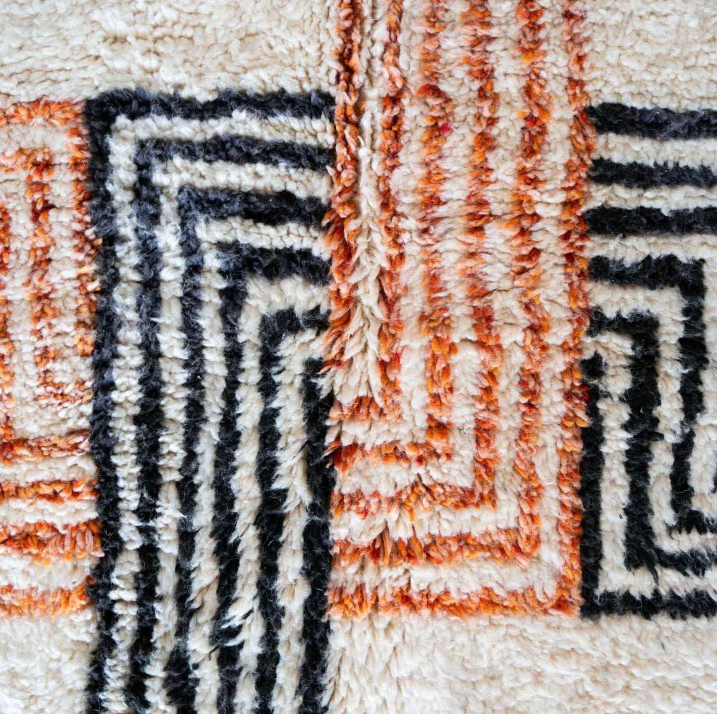 Artisanal handmade wool rug with vibrant orange and black geometric cross patterns on a soft white base, accented by intricate fringed edges, ideal for modern and eclectic decor.