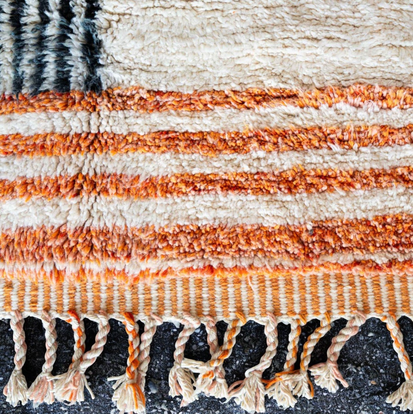 Artisanal handmade wool rug with vibrant orange and black geometric cross patterns on a soft white base, accented by intricate fringed edges, ideal for modern and eclectic decor.