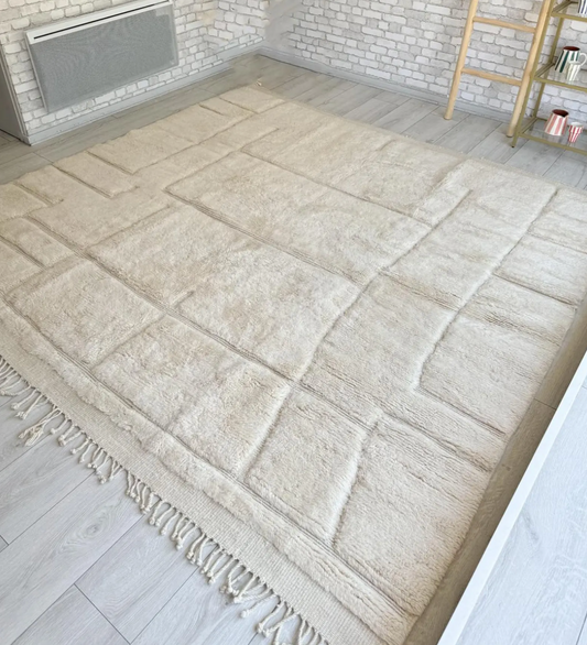 Handmade Moroccan cream Beni Ourain-style wool rug with a minimalist geometric pattern, displayed in a bright, modern room with a light wood floor, a decorative shelf with colorful mugs, a potted plant, and natural light creating a calm and cozy atmosphere