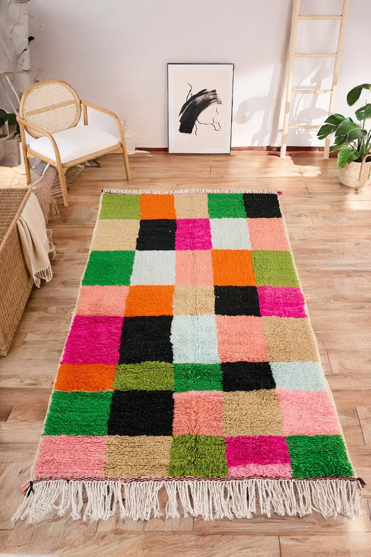 Handmade Moroccan wool rug featuring a bold multicolor checkerboard design with vibrant shades of pink, green, orange, black, beige, and blue, displayed on a herringbone wooden floor in a modern boho room with a rattan chair, potted plant, and minimalist wall art