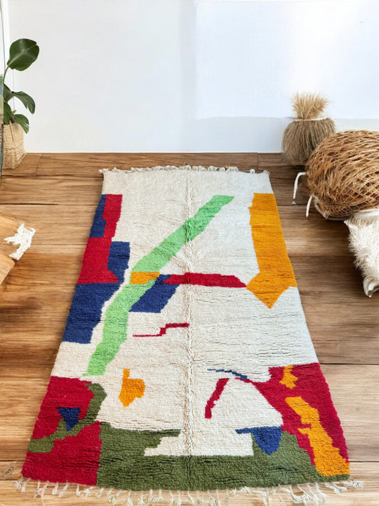 Handmade Moroccan wool rug featuring a vibrant abstract design in bold colors including red, green, blue, orange, and cream, displayed on a light wood floor in a modern boho-style room with a decorative shelf, potted plant, and natural light