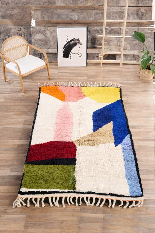 Handmade Moroccan wool rug featuring a vibrant abstract geometric design in bold colors like orange, pink, yellow, blue, green, red, and black, placed on a wooden floor in a boho-style room with a rattan chair, potted plant, and modern black-and-white wall art