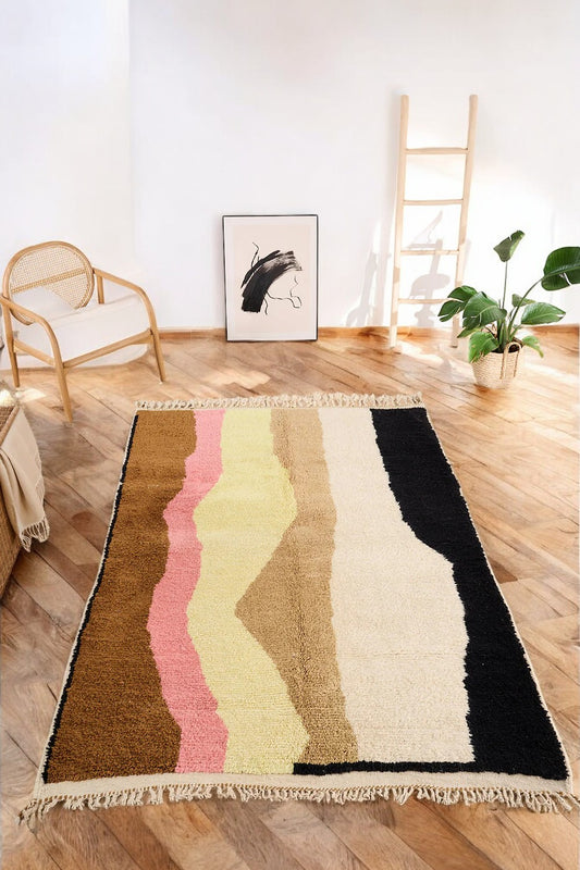 Handmade Moroccan wool rug featuring an abstract wave design with earthy tones of brown, beige, black, and pastel shades of pink and yellow, placed on a herringbone wooden floor in a modern boho-style room with a rattan chair, potted plant, and minimalist wall art.