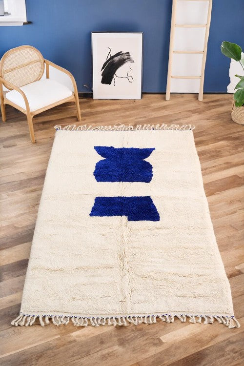 Handmade Moroccan wool rug in a minimalist cream color featuring a bold blue abstract design, displayed on a herringbone wooden floor in a modern boho room with a natural rattan chair, potted plant, and black-and-white wall art for a serene and stylish look