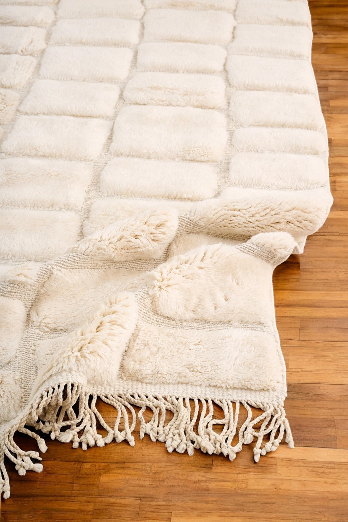Handmade Moroccan wool rug in a soft cream color featuring a textured square pattern, displayed on a wooden floor in a bright minimalist room with a rattan chair, potted plant, and black-and-white abstract wall art.
