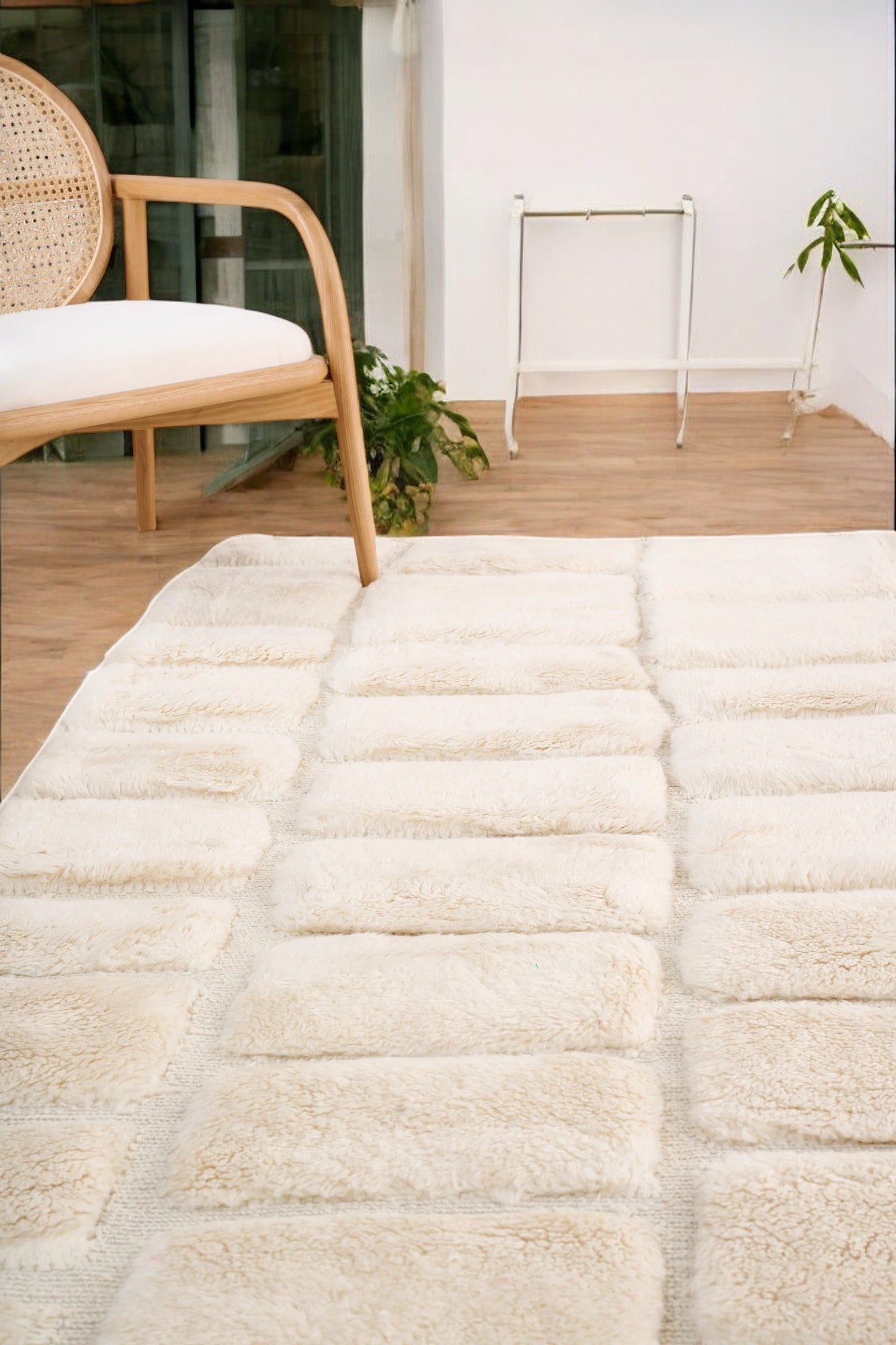 Handmade Moroccan wool rug in a soft cream color featuring a textured square pattern, displayed on a wooden floor in a bright minimalist room with a rattan chair, potted plant, and black-and-white abstract wall art.
