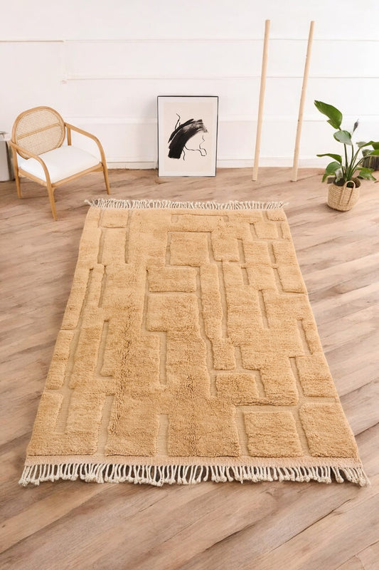 Handmade Moroccan wool rug in a warm beige color with a geometric textured pattern, displayed on a wooden floor in a modern minimalist room featuring a rattan chair, potted plant, and abstract black-and-white wall art