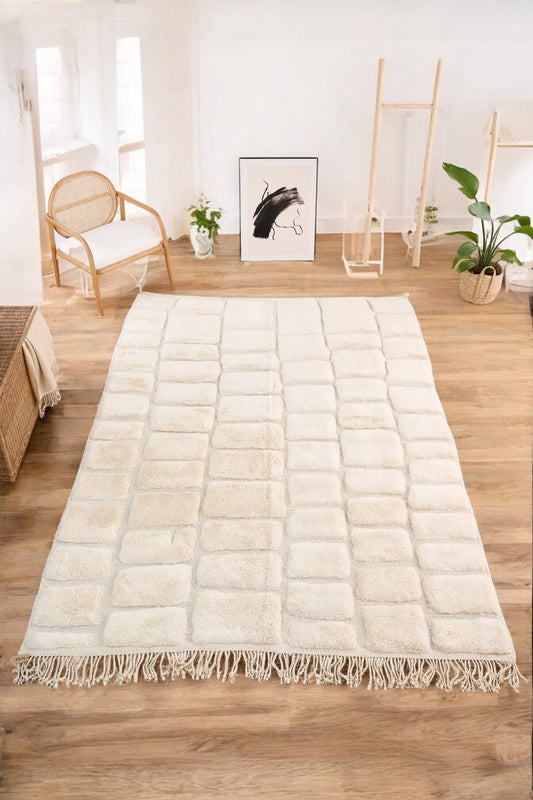 Handmade Moroccan wool rug in a soft cream color featuring a textured square pattern, displayed on a wooden floor in a bright minimalist room with a rattan chair, potted plant, and black-and-white abstract wall art.
