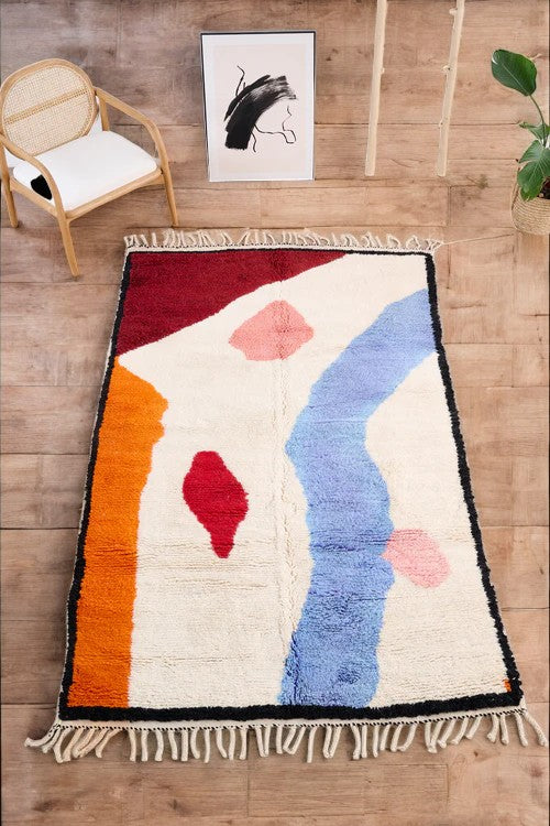 Handmade Moroccan wool rug with a bold abstract geometric design featuring vibrant colors like orange, pink, yellow, blue, green, red, and black, placed on a herringbone wooden floor in a modern boho room with a rattan chair, potted plant, and minimal wall art.