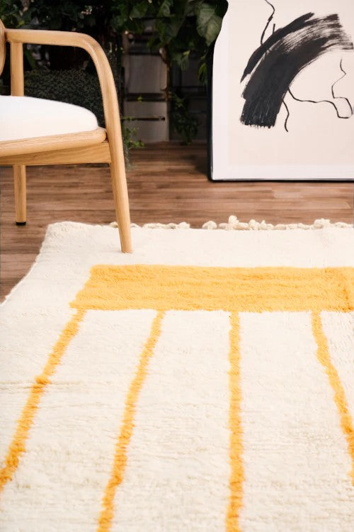 Handmade Moroccan wool rug with a minimalist cream base featuring vibrant yellow vertical stripes and a bold red border, styled on a wooden floor in a cozy boho room with a rattan chair, potted plant, and black-and-white abstract wall art