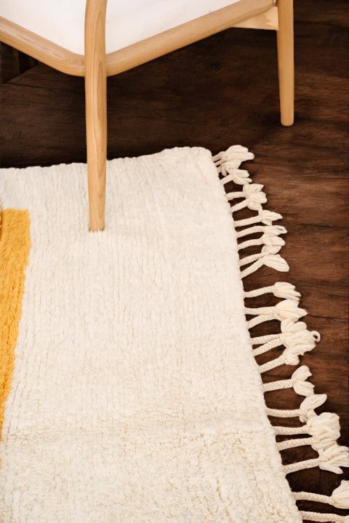 Handmade Moroccan wool rug with a minimalist cream base featuring vibrant yellow vertical stripes and a bold red border, styled on a wooden floor in a cozy boho room with a rattan chair, potted plant, and black-and-white abstract wall art