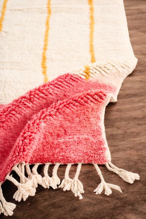 Handmade Moroccan wool rug with a minimalist cream base featuring vibrant yellow vertical stripes and a bold red border, styled on a wooden floor in a cozy boho room with a rattan chair, potted plant, and black-and-white abstract wall art