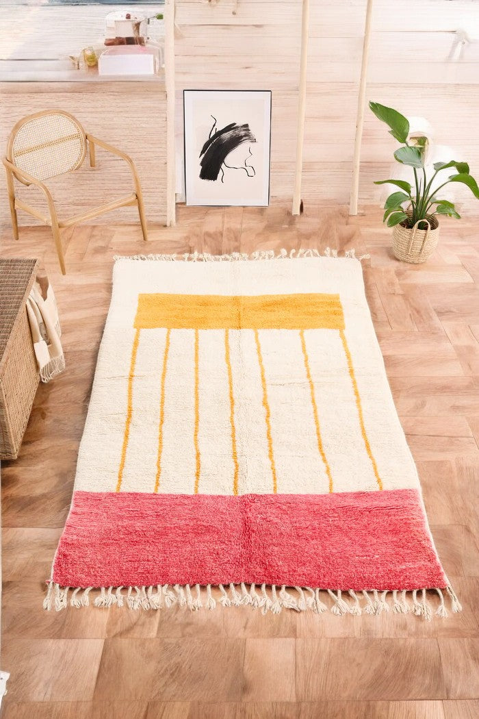 Handmade Moroccan wool rug with a minimalist cream base featuring vibrant yellow vertical stripes and a bold red border, styled on a wooden floor in a cozy boho room with a rattan chair, potted plant, and black-and-white abstract wall art